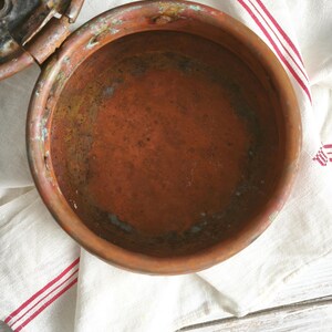 Puede incluir: Un recipiente redondo de cobre desgastado con tapa abatible. El interior muestra una p&aacute;tina de color marr&oacute;n rojizo profundo con oxidaci&oacute;n visible y manchas de cardenillo verde. La superficie met&aacute;lica parece envejecida y texturizada.