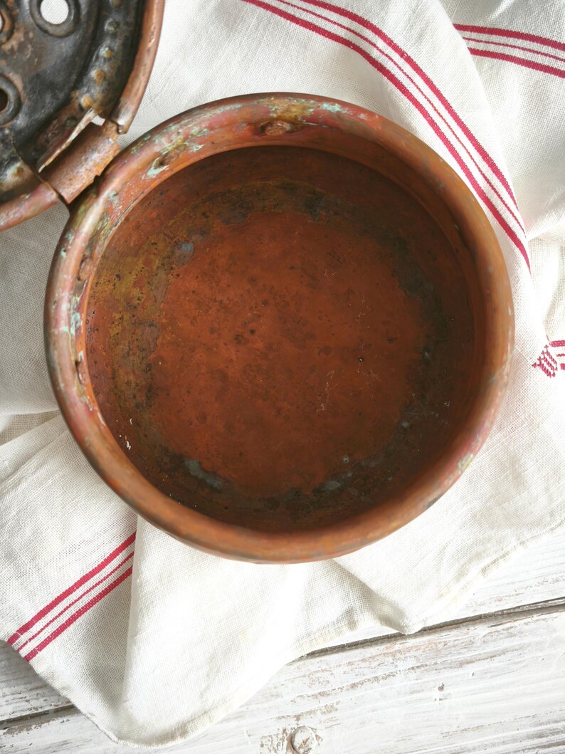 Puede incluir: Un recipiente redondo de cobre desgastado con tapa abatible. El interior muestra una p&aacute;tina de color marr&oacute;n rojizo profundo con oxidaci&oacute;n visible y manchas de cardenillo verde. La superficie met&aacute;lica parece envejecida y texturizada.
