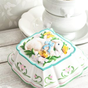 Könnte beinhalten: Eine weiße Keramikschale in Lammform mit einer blauen Schleife, umgeben von Blumen und Schmetterlingen. Die Schale hat einen gewellten Rand mit grünen und rosa Akzenten, ideal für Ostern oder Frühlingsdekoration.