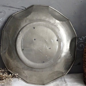 May include: A silver-colored, octagonal metal plate with a slightly raised rim. The plate has a smooth, slightly textured surface and five small holes in the center.