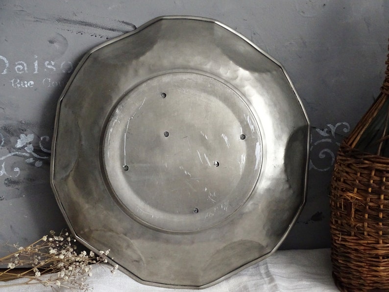 May include: A silver-colored, octagonal metal plate with a slightly raised rim. The plate has a smooth, slightly textured surface and five small holes in the center.