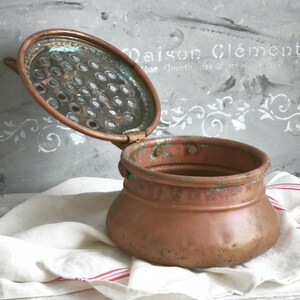 Puede incluir: Una olla redonda de cobre con una tapa perforada con bisagras. El metal muestra signos de oxidaci&oacute;n con manchas de p&aacute;tina verde en el borde y en la parte inferior de la tapa. La olla tiene un cuerpo ancho y curvo con un acabado desgastado.