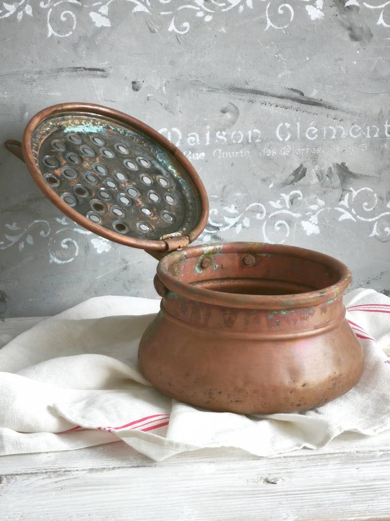 Puede incluir: Una olla redonda de cobre con una tapa perforada con bisagras. El metal muestra signos de oxidaci&oacute;n con manchas de p&aacute;tina verde en el borde y en la parte inferior de la tapa. La olla tiene un cuerpo ancho y curvo con un acabado desgastado.
