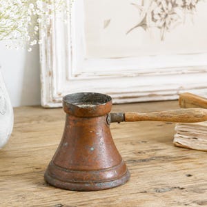 May include: A weathered copper coffee pot with a wide, tapered base and a narrow rim. The pot features a long, light-colored wooden handle attached with a metal rivet. The copper surface shows signs of oxidation with small green and dark spots.