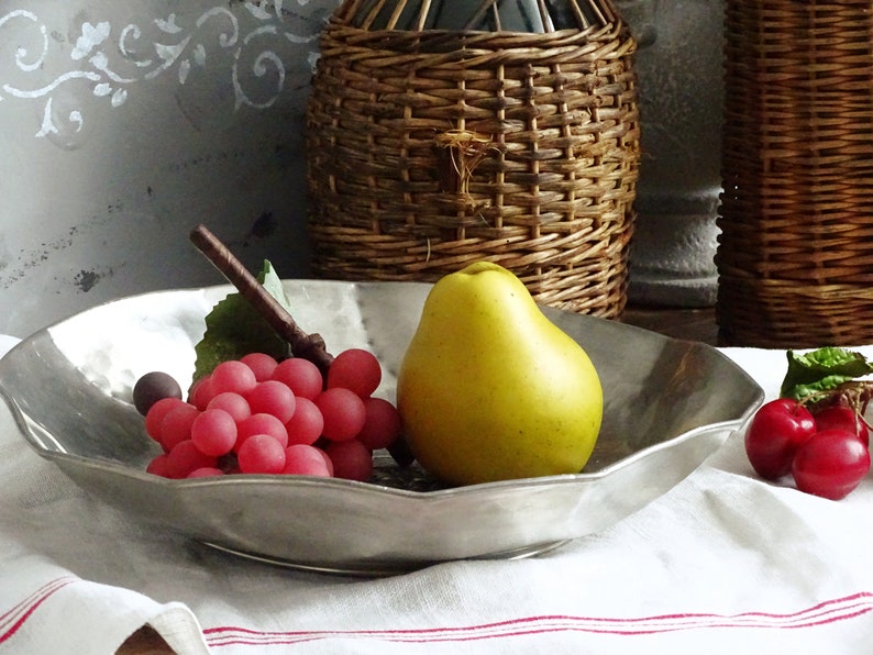 May include: A silver serving bowl with a scalloped edge, filled with red grapes, a yellow pear, and red cherries. The bowl is sitting on a white tablecloth with red stripes.
