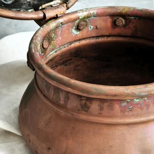 Puede incluir: Una olla de cobre desgastada con una tapa perforada con bisagras. La superficie met&aacute;lica muestra un tono marr&oacute;n rojizo c&aacute;lido con signos visibles de oxidaci&oacute;n, incluyendo parches de p&aacute;tina verde y manchas oscuras. La tapa tiene agujeros circulares.
