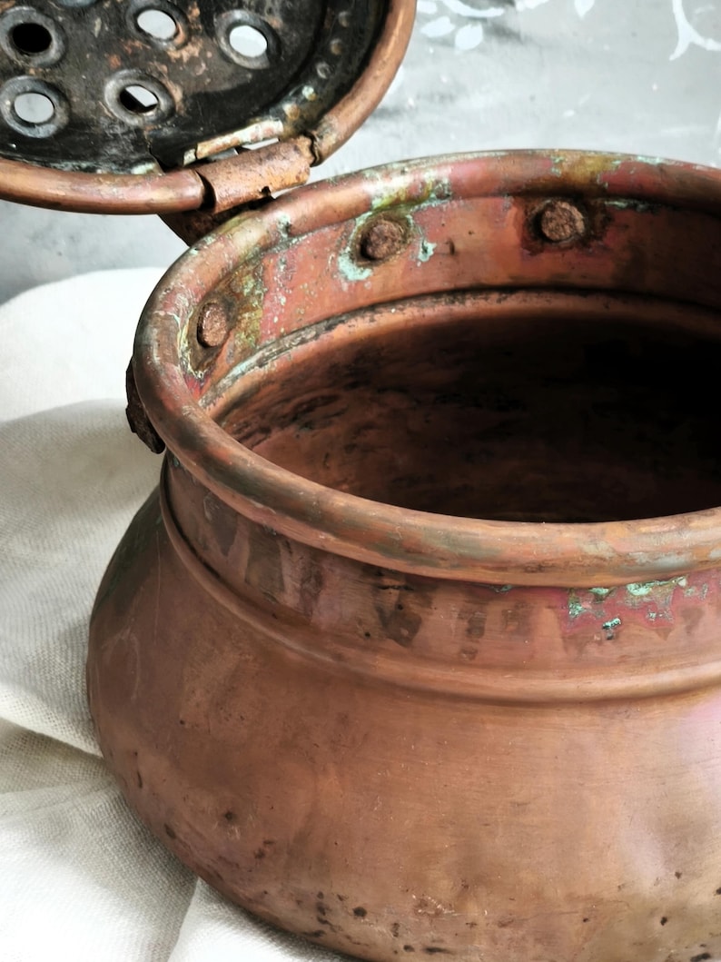 Puede incluir: Una olla de cobre desgastada con una tapa perforada con bisagras. La superficie met&aacute;lica muestra un tono marr&oacute;n rojizo c&aacute;lido con signos visibles de oxidaci&oacute;n, incluyendo parches de p&aacute;tina verde y manchas oscuras. La tapa tiene agujeros circulares.