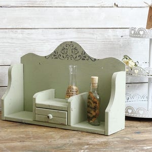 May include: A distressed, sage green wooden shelf with a small drawer and decorative top. Two glass bottles with cork stoppers sit on the shelf. A white tiered stand with heart cutouts is in the background.