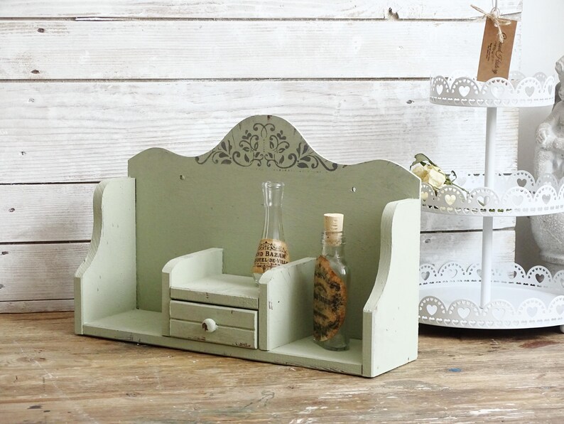 May include: A distressed, sage green wooden shelf with a small drawer and decorative top. Two glass bottles with cork stoppers sit on the shelf. A white tiered stand with heart cutouts is in the background.