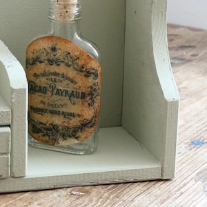 May include: A clear glass bottle with a cork stopper and a vintage label, displayed on a light green wooden shelf. The label features ornate text and designs. The shelf is part of a larger, aged wooden structure.