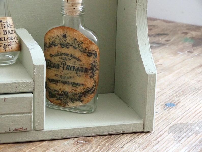May include: A clear glass bottle with a cork stopper and a vintage label, displayed on a light green wooden shelf. The label features ornate text and designs. The shelf is part of a larger, aged wooden structure.