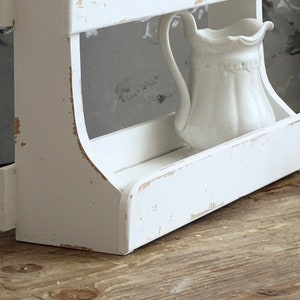 May include: A white wooden wall shelf with a distressed finish featuring visible wood grain and worn edges. The shelf has two tiers and holds a small, white ceramic pitcher with a scalloped rim and a curved handle on the bottom shelf surface.