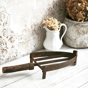 May include: An antique, rusty metal trivet with a wooden handle. The trivet has a triangular shape with three parallel bars and is set on a white wooden surface. A white pitcher with dried flowers and a decorative pot are in the background.