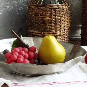 May include: A silver serving bowl with a scalloped edge, filled with red grapes, a yellow pear, and red cherries. The bowl is sitting on a white tablecloth with red stripes.