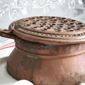 Puede incluir: Un recipiente redondo de cobre con una tapa plana perforada que presenta numerosos orificios circulares. La superficie de cobre muestra signos de oxidaci&oacute;n con manchas de p&aacute;tina verde y un acabado texturizado y desgastado. Tiene un asa met&aacute;lica lateral.