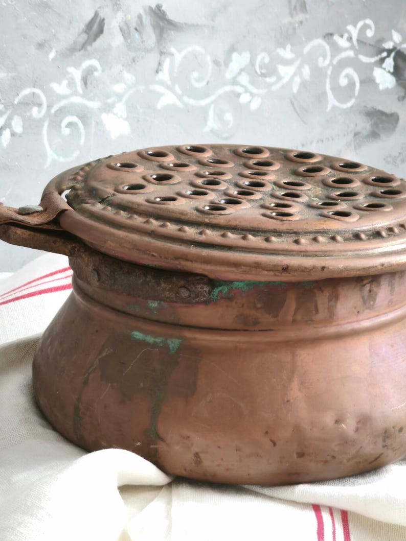Puede incluir: Un recipiente redondo de cobre con una tapa plana perforada que presenta numerosos orificios circulares. La superficie de cobre muestra signos de oxidaci&oacute;n con manchas de p&aacute;tina verde y un acabado texturizado y desgastado. Tiene un asa met&aacute;lica lateral.
