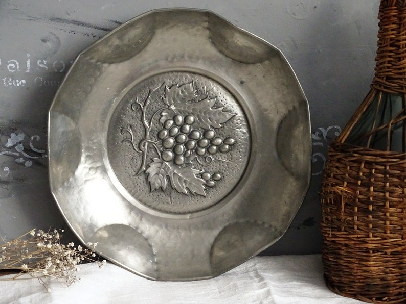 May include: A silver-toned metal plate with a raised design of a grapevine and grapes. The plate has a scalloped edge and a hammered finish.