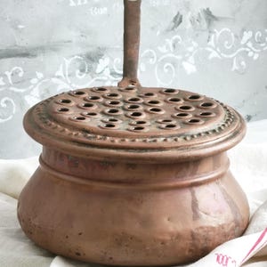 Puede incluir: Un recipiente redondo de cobre con una base ancha y una tapa extra&iacute;ble que presenta numerosas perforaciones circulares. La tapa tiene un asa vertical central y la superficie de cobre muestra una p&aacute;tina envejecida con oxidaci&oacute;n.