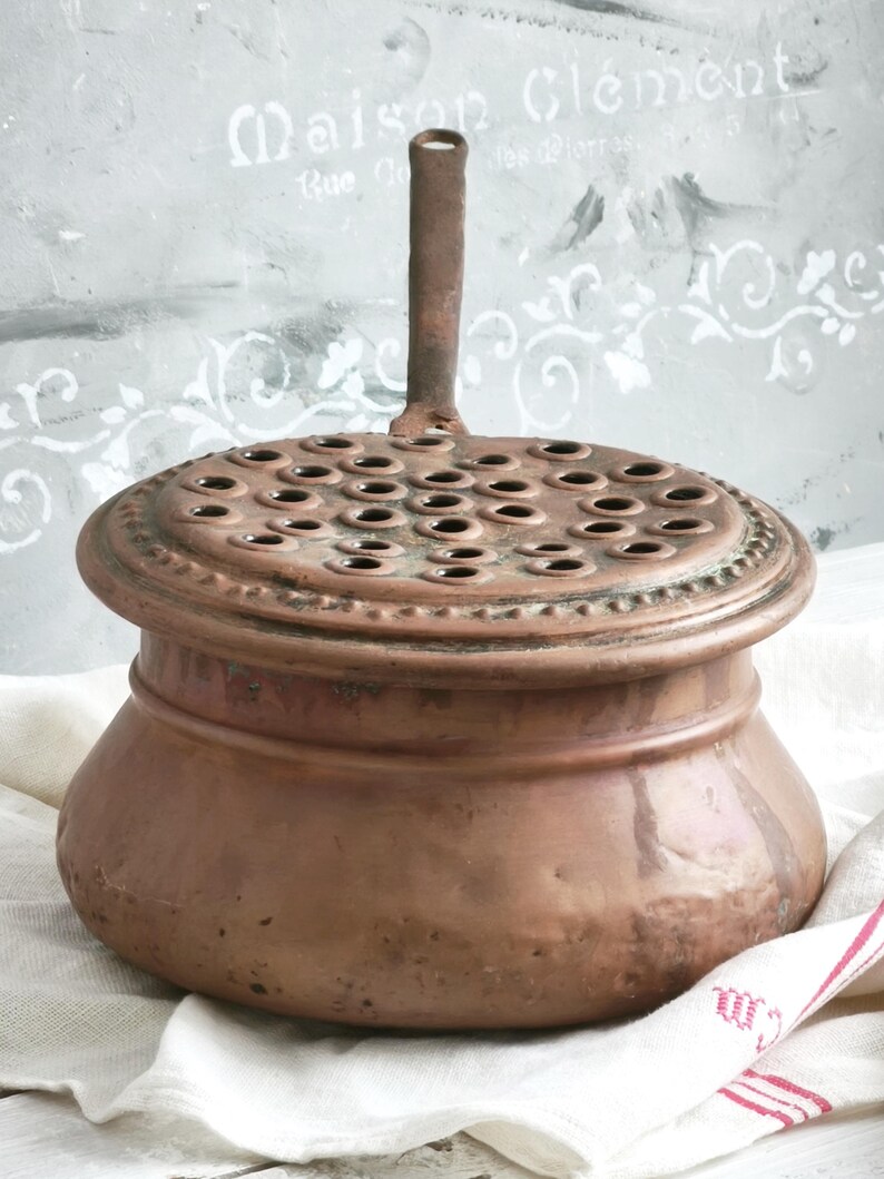Puede incluir: Un recipiente redondo de cobre con una base ancha y una tapa extra&iacute;ble que presenta numerosas perforaciones circulares. La tapa tiene un asa vertical central y la superficie de cobre muestra una p&aacute;tina envejecida con oxidaci&oacute;n.