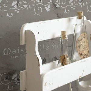 May include: A white wooden wall shelf with a distressed finish, featuring two clear glass bottles with cork stoppers. The taller bottle has a beige oval label with text, and the smaller bottle has a smaller beige label. A white ceramic pitcher is visible below.