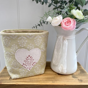 May include: A light green fabric basket with a heart applique and a white pitcher with flowers. The basket has a floral pattern and a heart-shaped patch with a decorative design. The white pitcher holds pink and white flowers.