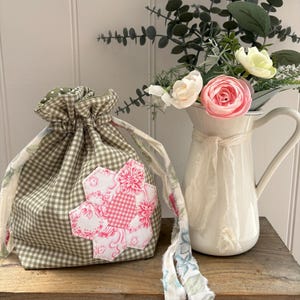 May include: A drawstring pouch made of sage green and white gingham fabric, featuring a central floral-patterned hexagon appliqué in shades of pink and white. The pouch has long, frayed-edge fabric ties with a light blue and green floral print.