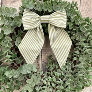 May include: A eucalyptus wreath with a large bow. The bow has a green and white striped pattern. The wreath is made of green eucalyptus leaves and is set against a wooden background.
