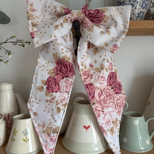 May include: A large fabric bow featuring a white background with a repeating pattern of pink and dark rose-coloured roses, green leaves, and small brown sprigs. The bow has two long, pointed tails hanging down from the centre knot.