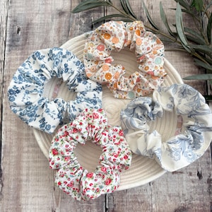 May include: Five fabric scrunchies with floral patterns in various colors, including blue, orange, and red. The scrunchies are arranged on a white woven plate, with a wooden background and greenery.