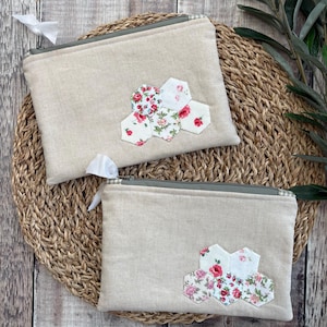 May include: Two rectangular beige linen zip pouches featuring a cluster of hexagonal fabric patches with floral patterns in shades of red, pink, and green. Each pouch has a green zip closure with a small white fabric pull tab attached.