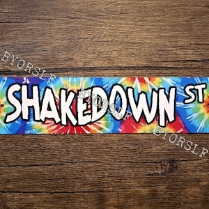 Könnte beinhalten: Ein farbenfrohes Batik-Schild mit dem Text "Shakedown St." in weißen Buchstaben.