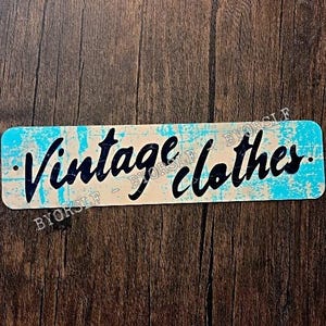 May include: A rectangular sign with the words "Vintage clothes" in a black, cursive font. The sign has a cream-colored background with a distressed, light blue border. The sign is placed on a dark brown wooden surface.