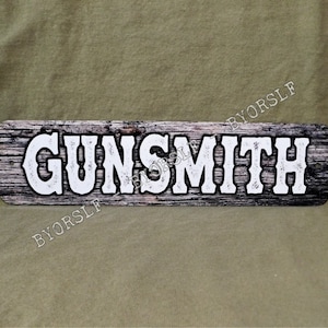May include: A rustic wooden sign with the word "GUNSMITH" printed in white letters. The sign has a distressed wood look and is perfect for a western-themed decor.