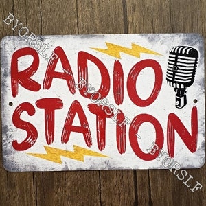 Peut inclure: Une pancarte en métal blanc avec du texte rouge qui dit "RADIO STATION" avec un graphique de microphone stylisé. La pancarte a des graphiques de foudre jaunes.