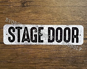 May include: A white rectangular sign with black text that reads "STAGE DOOR". The sign has a distressed, vintage look.