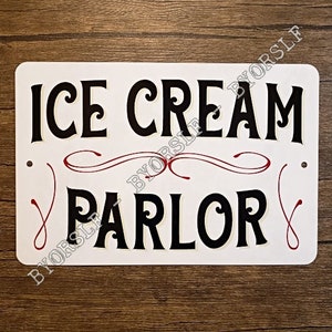 Op de afbeelding: Een wit metalen bord met zwarte tekst die "ICE CREAM PARLOR" luidt met rode en witte decoratieve krulletjes.