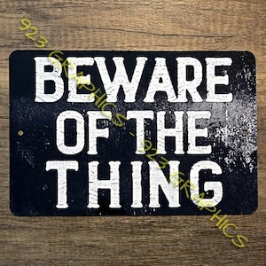 以下が含まれることがあります： 「BEWARE OF THE THING」の文字が大きく白く書かれた、長方形の黒い金属製の看板。 ヴィンテージ感のある、使い古された外観で、摩耗や損傷が見られます。