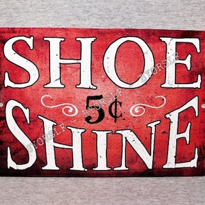 May include: Red metal sign with white text that reads "Shoe Shine 5¢". The sign has a distressed, vintage look.