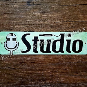 Puede incluir: Un letrero de metal verde claro con la palabra "Studio" en letras negras. Un micrófono y un cable se representan en el lado izquierdo del letrero.