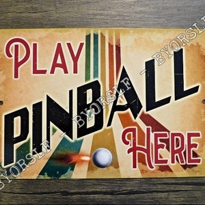 May include: A vintage-style metal sign with a retro design. It says "Play Pinball Here" in bold, black letters with red accents. The background is a faded yellow with colourful stripes and a pinball.