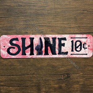May include: A rectangular metal sign with the word "SHINE" in large black letters. The letter "I" is replaced with a shoe graphic. The sign has a pink and white background and includes the text "10¢".