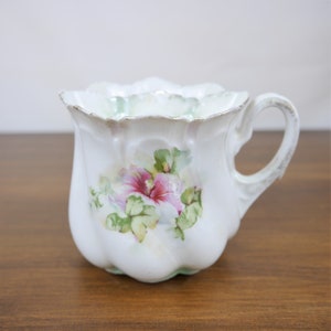 May include: White porcelain teacup with a scalloped rim and handle. The cup features a hand-painted floral design with pink and green accents. The handle and rim have gold trim. The cup is sitting on a wooden surface.