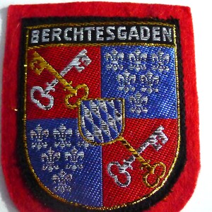 Peut inclure: Écusson en forme de bouclier avec le mot "BERCHTESGADEN" en haut. Le bouclier est divisé en quadrants rouges et bleus, avec des clés dorées et des motifs de fleur de lys blancs. L'écusson a une bordure en feutre rouge.