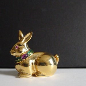 May include: A gold rabbit figurine with green and purple enamel accents. The rabbit has pink eyes and is sitting on a white surface.