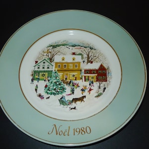May include: A vintage Christmas plate with a green border and a gold rim. The centre features a snowy village scene with houses, a Christmas tree, and people. The text "Noël 1980" is printed in gold on the bottom.