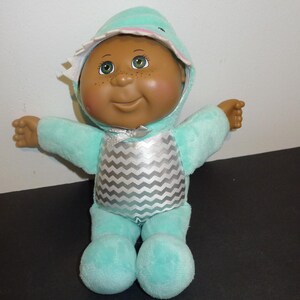 Peut inclure: Poupée Cabbage Patch Kids vêtue d'une tenue à capuche turquoise avec un motif chevron argenté sur la poitrine. La poupée a la peau brune, les yeux verts et des taches de rousseur. La capuche a une bordure blanche.