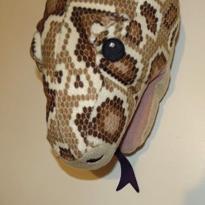 May include: A soft toy snake head featuring a detailed pattern of brown, beige, and cream scales. The snake has a black eye and a purple tongue. The plush toy is designed to be cuddly.