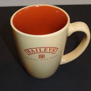 Peut inclure: Une tasse en céramique beige avec un intérieur orange. La tasse porte l'inscription "BAILEYS" "ORIGINAL IRISH CREAM" en rouge sur le devant.