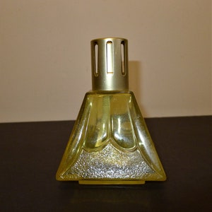 May include: A clear glass oil burner with a gold metal top and a textured surface. The burner is shaped like a pyramid with a rounded top.