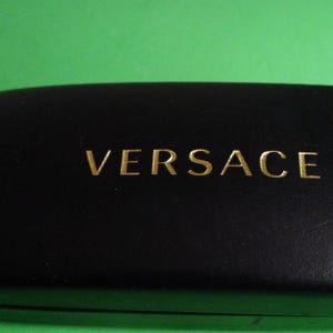 May include: Black rectangular glasses case with the gold-coloured word "VERSACE" centred on the top. The case has a smooth, slightly rounded shape and is set against a green background.